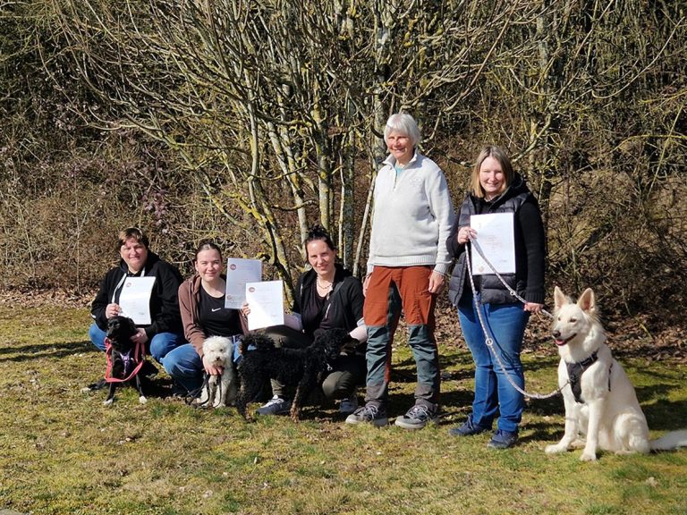 Sechs Personen stehen mit Hunden vor einem Baum und halten Unterlagen in der Hand.